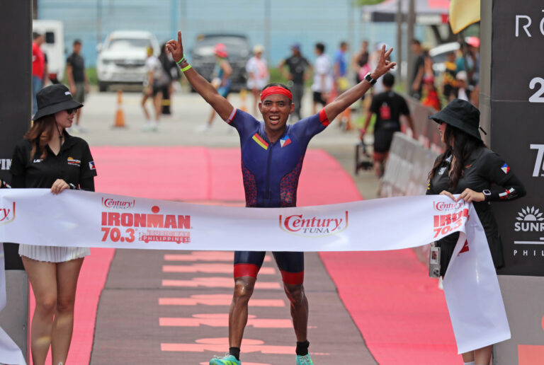 August Benedicto crossing a triathlon finish line with his arms raised