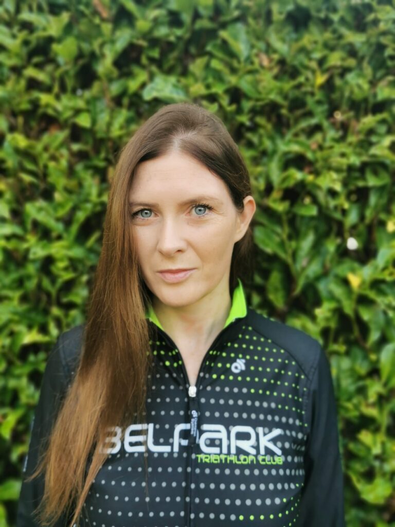 Andrea Brennan, Endurance Lab triathlete, in a Belpark Triathlon Club jacket in front of a green hedge